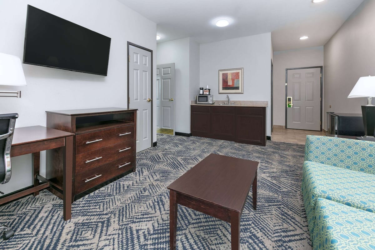 A hotel room with a flat-screen TV, a wooden desk, a patterned carpet, a sofa, and a kitchenette area.