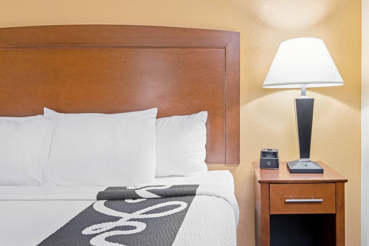 Una cama bien tendida con almohadas blancas y una colcha gris y blanca, una mesita de noche de madera con una lámpara y un reloj, y un cabecero de madera.