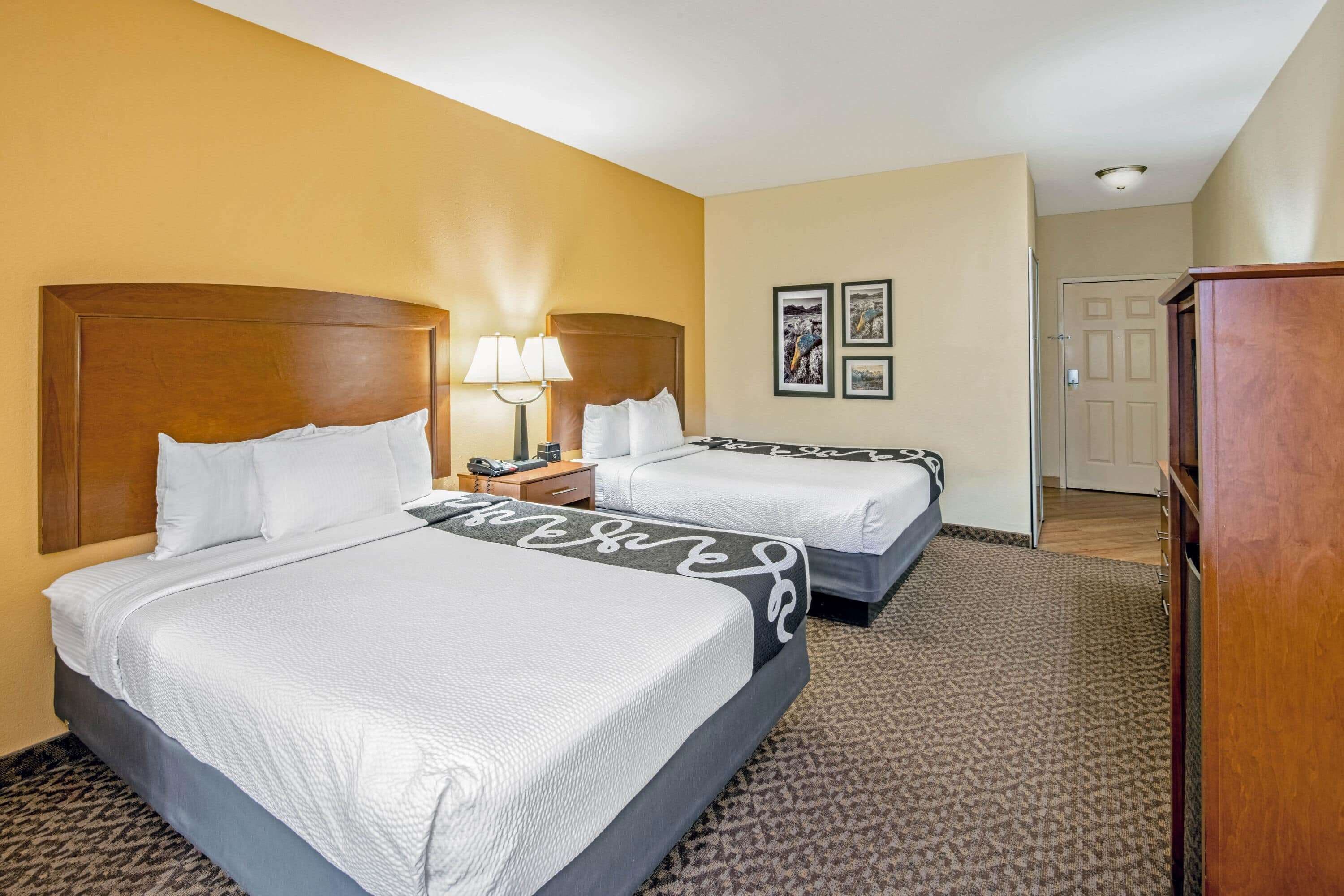 A hotel room with two beds, a nightstand, a lamp, and framed pictures on the wall.