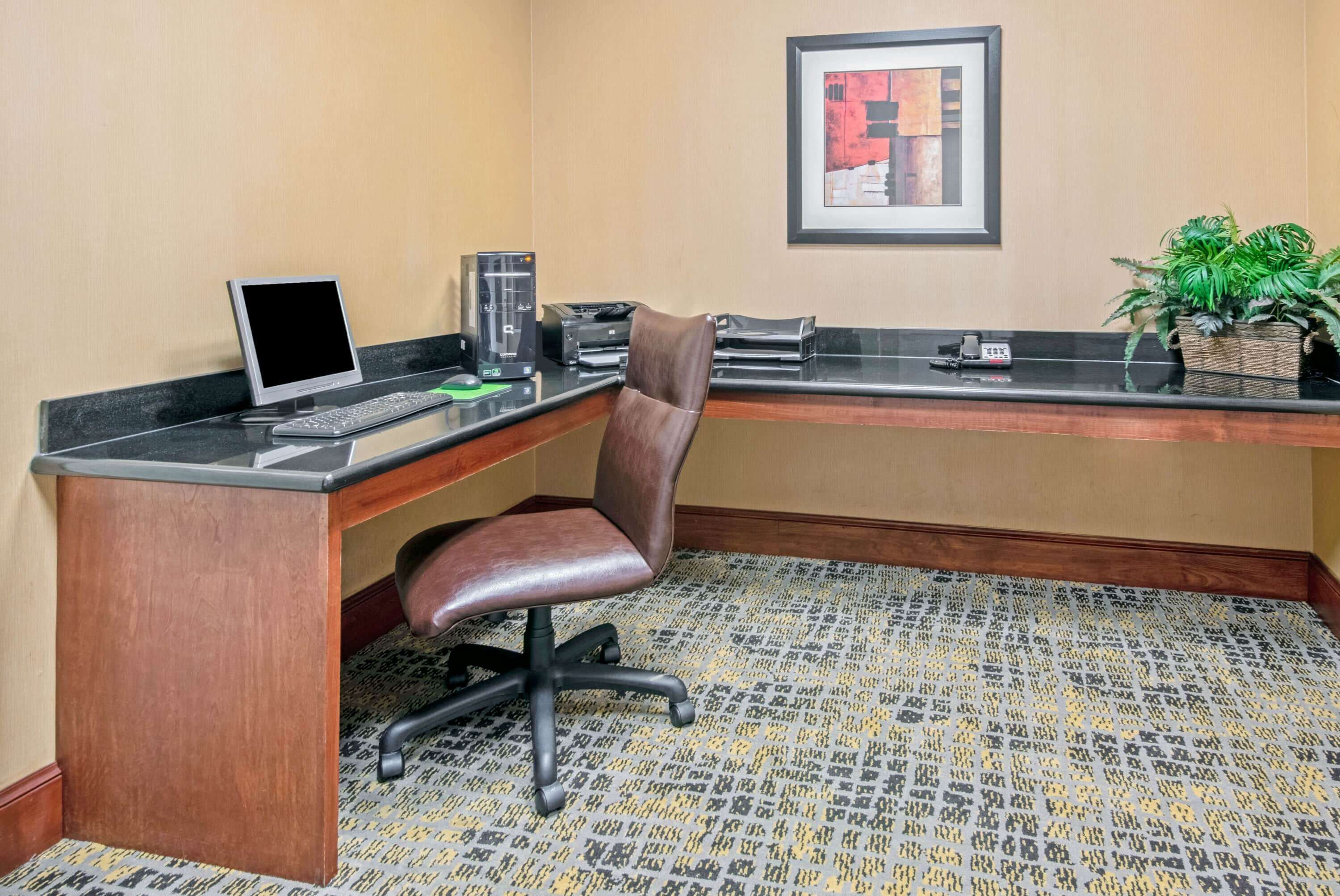 A hotel reception desk with a computer, printer, and phone. There is a framed picture on the wall and a plant on the counter.