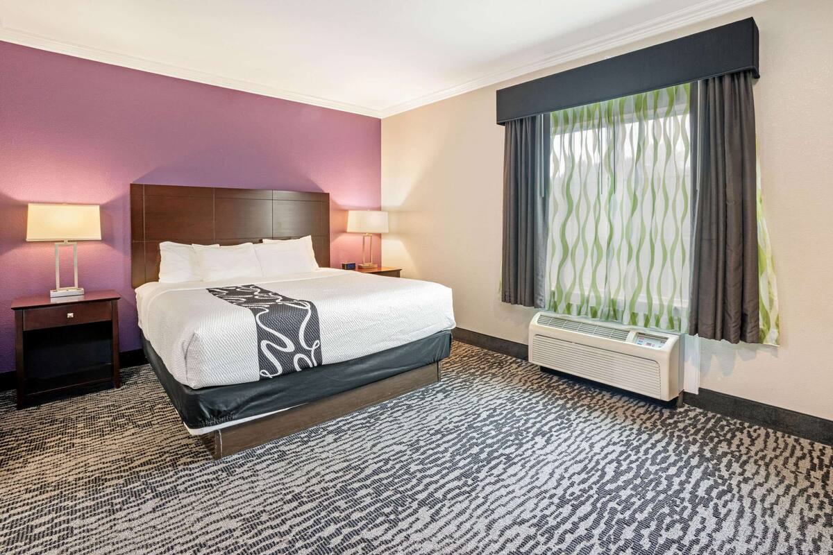 A hotel room with a large bed, two bedside tables with lamps, a window with curtains, and a wall-mounted air conditioner.