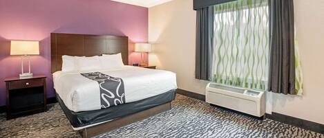 A hotel room with a large bed, two bedside tables with lamps, a window with curtains, and a wall-mounted air conditioner.