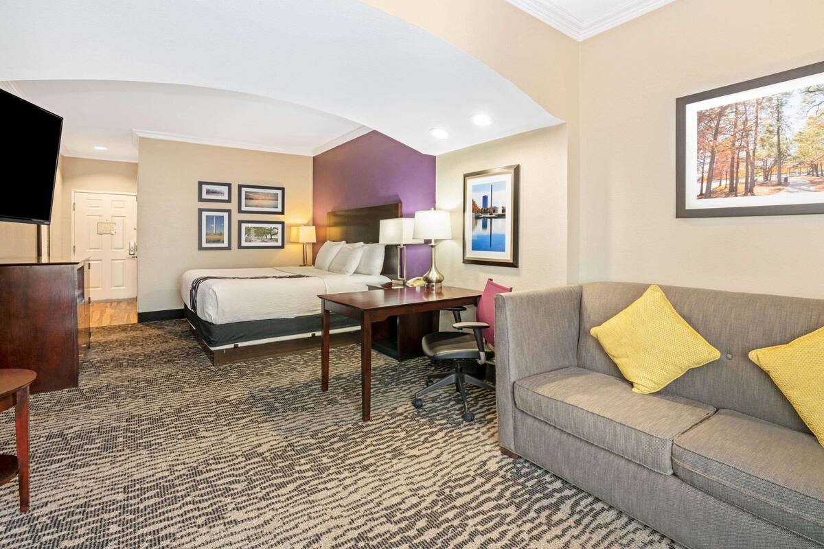 A hotel room with a bed, a gray sofa with yellow cushions, a small table, and framed pictures on the wall.