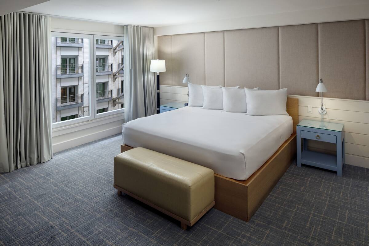A hotel room with a large bed, a bench, two bedside tables, and a view of the cityscape through the window.