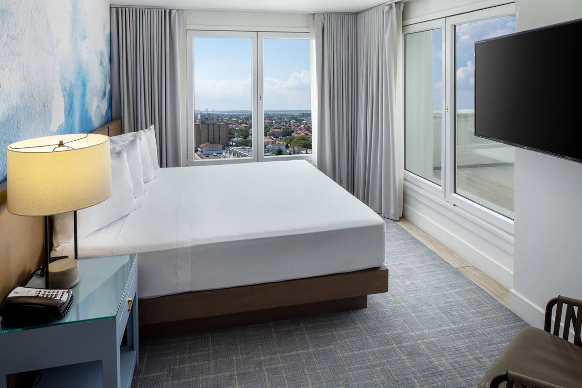 A hotel room with a large bed, a desk with a lamp, a flat-screen TV, and a view of the city through large windows.