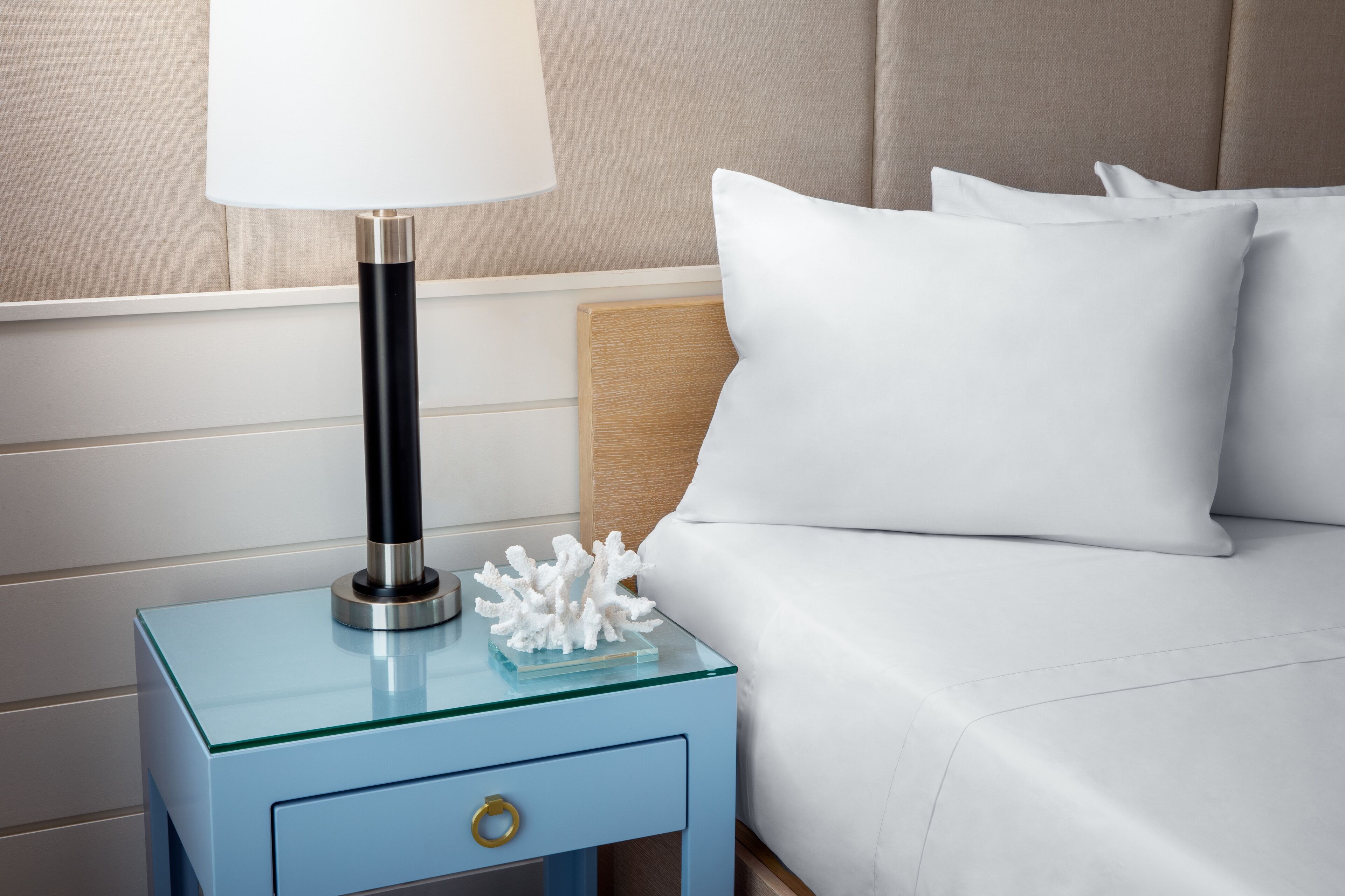 A neatly made bed with white linens, a bedside lamp, and a decorative coral piece on a glass nightstand.