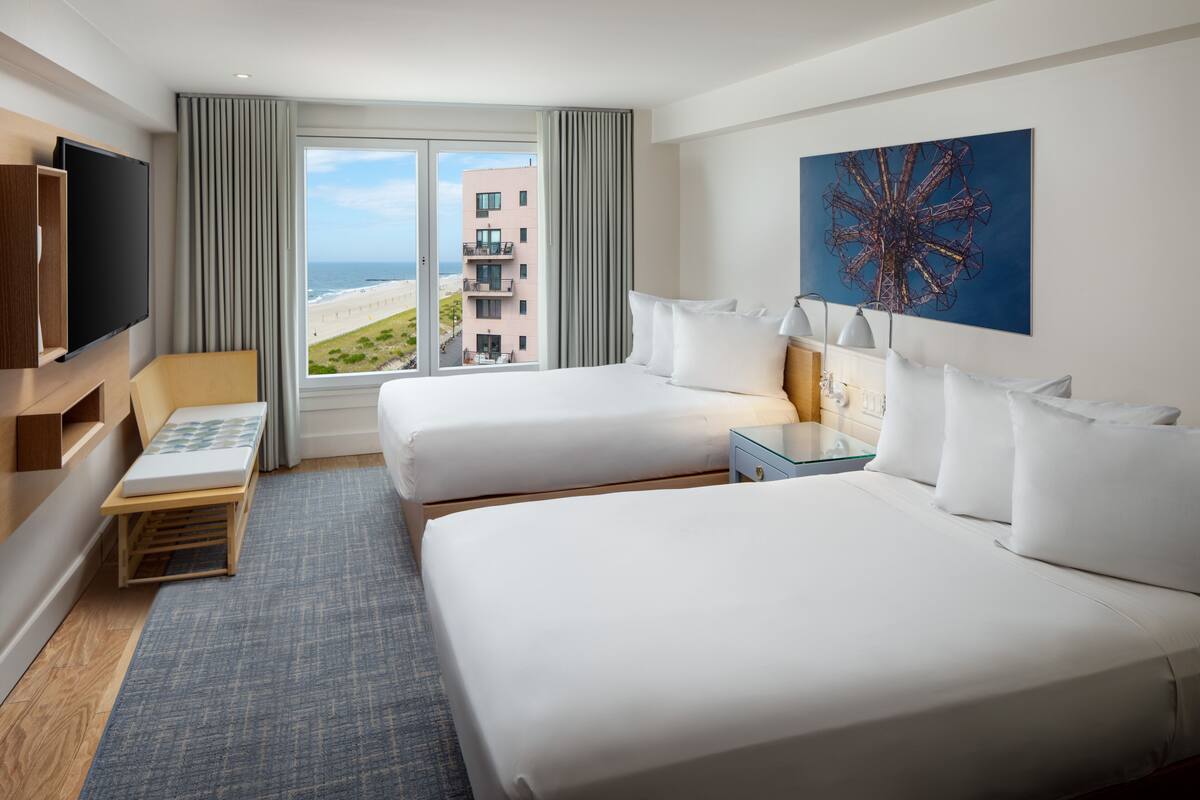 Habitación de hotel con dos camas, televisión, escritorio y ventana grande con vistas a la playa y edificios.