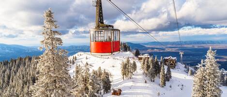 A red cable car ascending a snowy mountain slope with a cabin, surrounded by snow-covered trees and a distant view of a town.