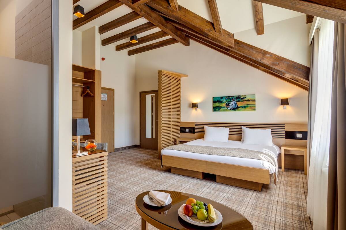 Una habitación de hotel con una cama grande, cabecera de madera, una mesita de noche con lámpara, una pequeña mesa con fruta y un cuadro en la pared.