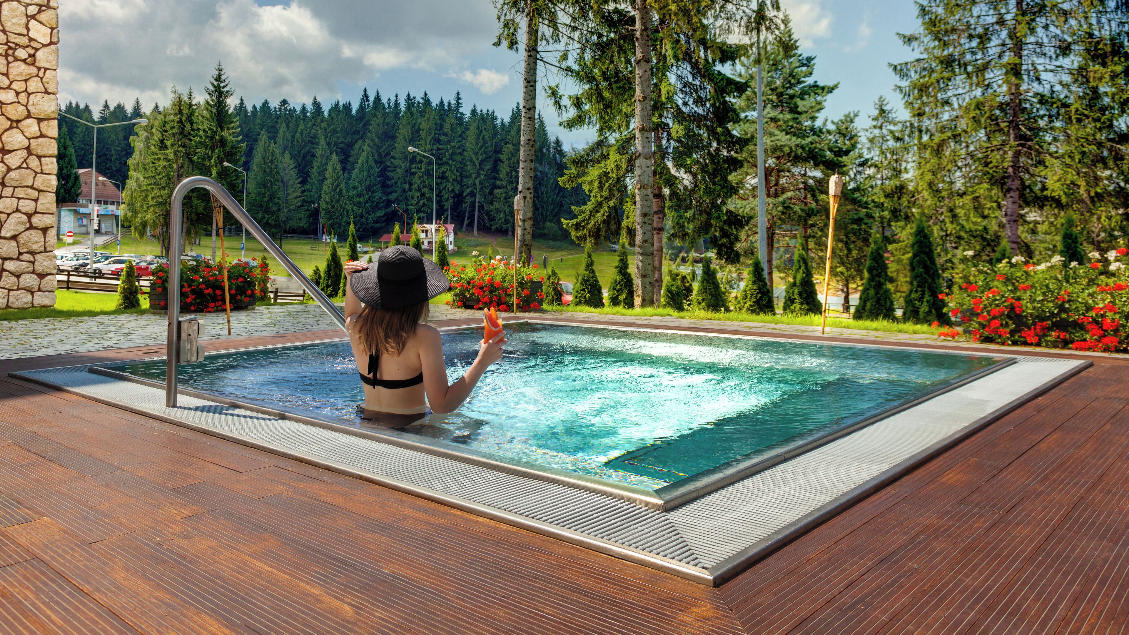 Une femme dans une piscine, avec une terrasse en bois, entourée d’arbres et de fleurs.
