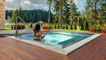 Une femme dans une piscine, avec une terrasse en bois, entourée d’arbres et de fleurs.