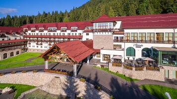 Traditional alpine-style hotel with a stone facade and red-tiled roof, set amidst a well-manicured lawn and stone wall, offering a mountain view.