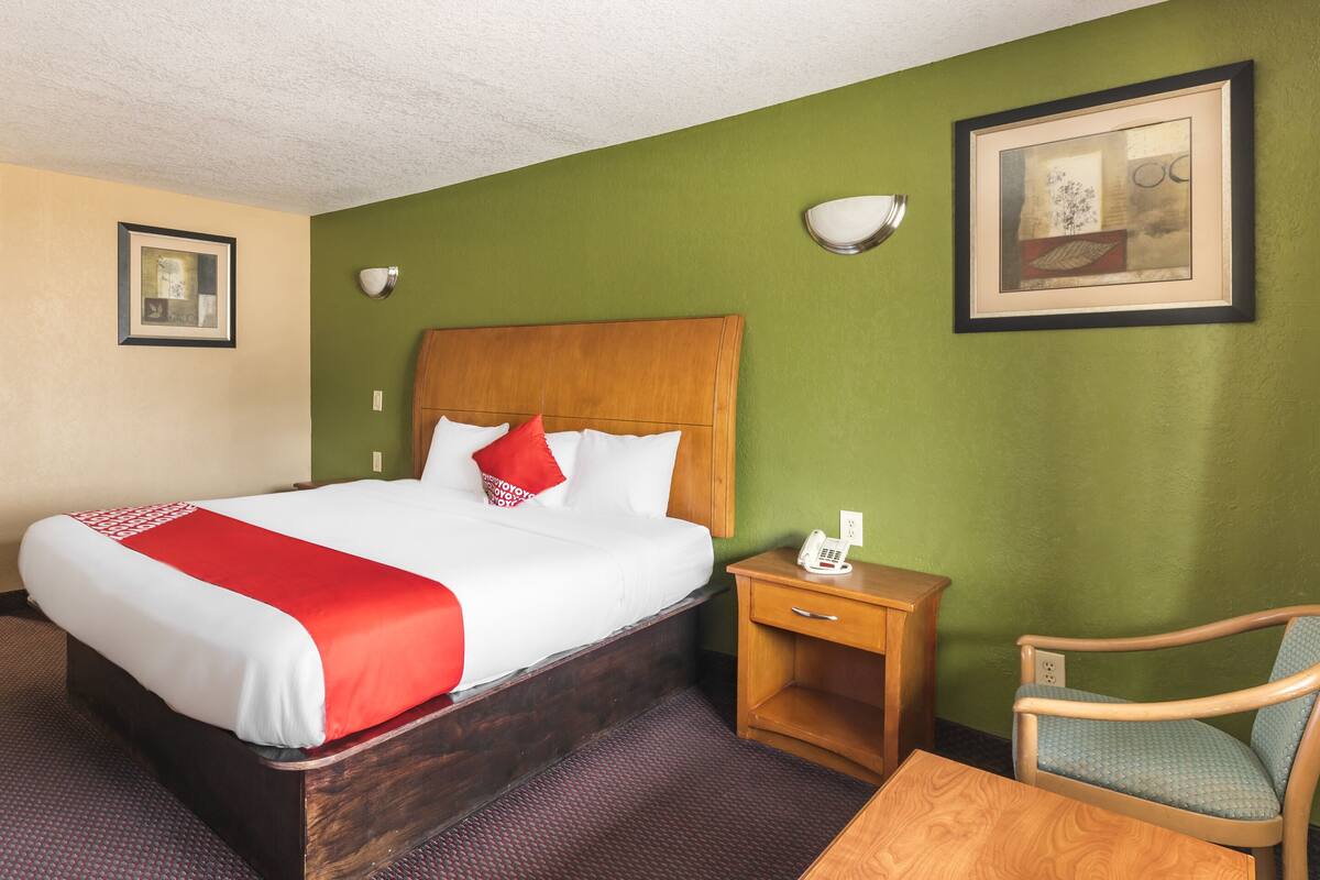 A hotel room with a wooden bed, a red and white bedspread, a wooden nightstand with a phone, a blue chair, and green walls.