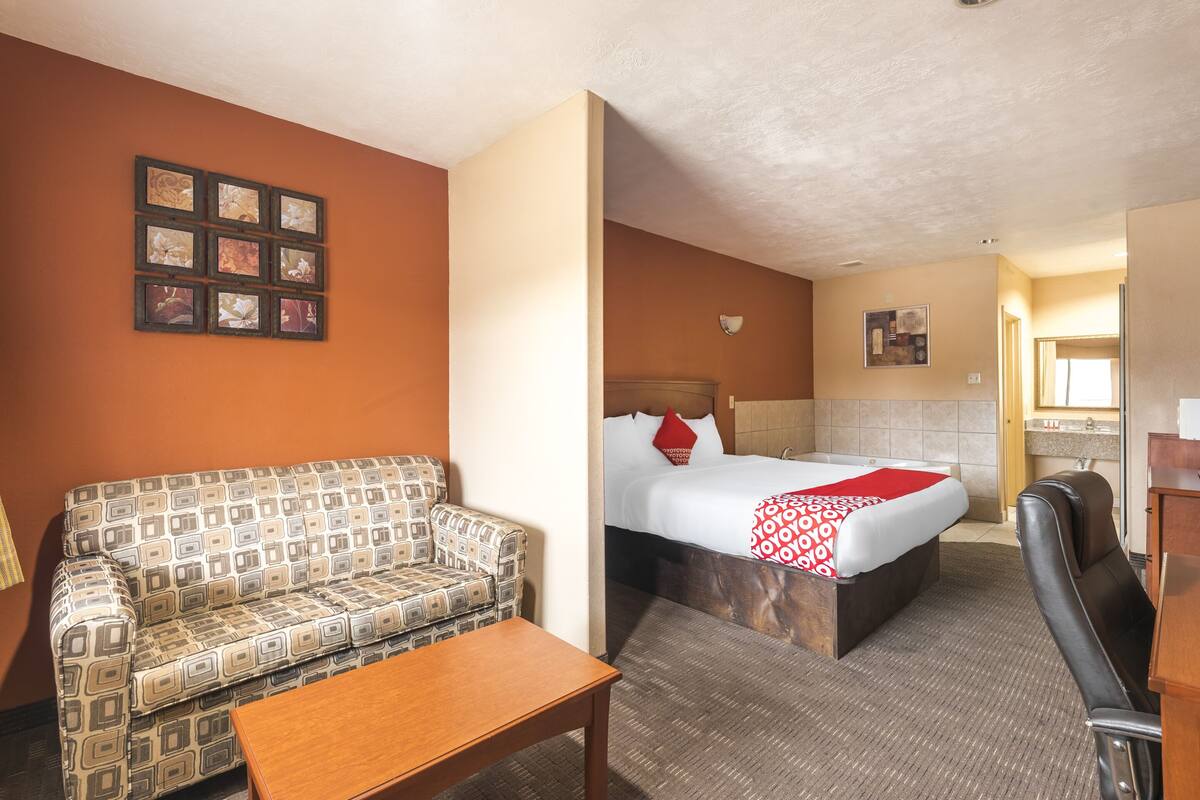 A hotel room with a patterned sofa, a wooden table, a bed with a red and white blanket, and a small desk with a chair.