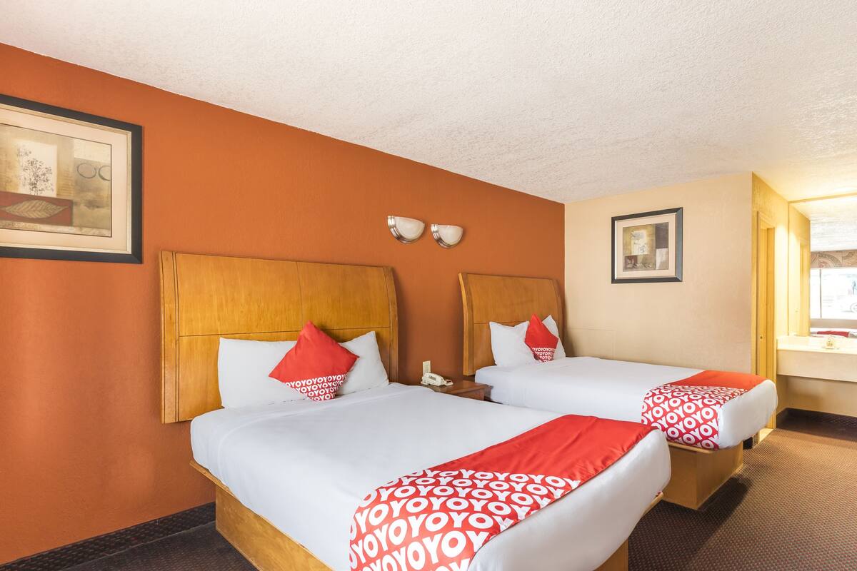 A hotel room with two beds, each with white bedding and red accents, a wooden headboard, a bedside table with a lamp, and a framed picture on the wall.