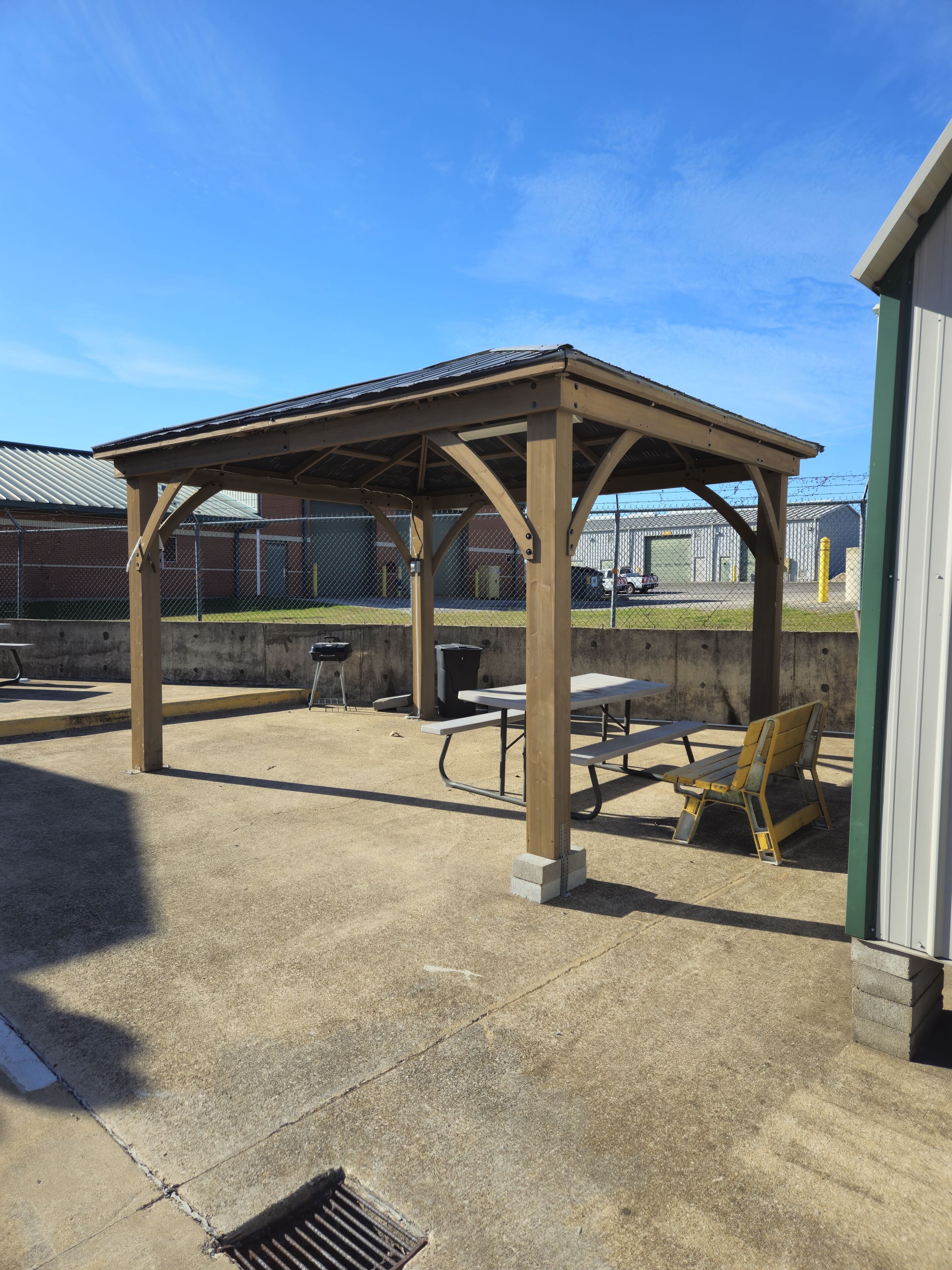 A covered outdoor seating area with picnic tables and benches, a concrete floor, and a metal fence in the background.