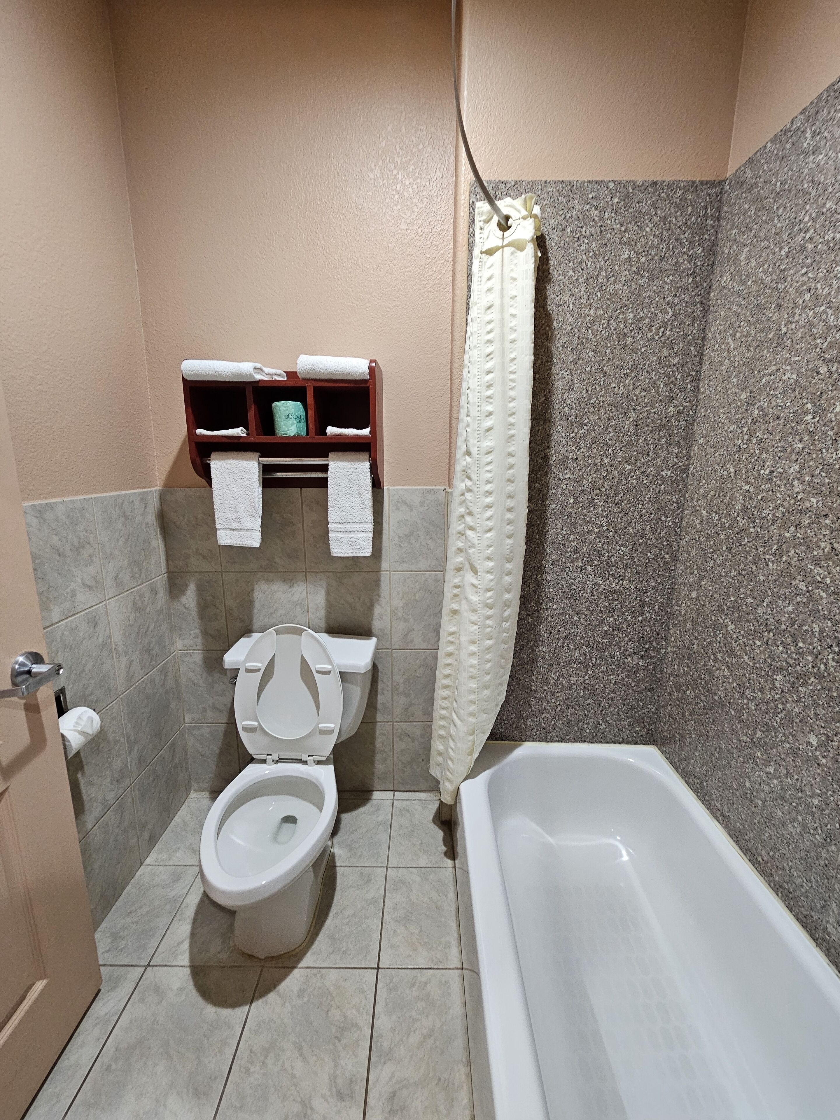 Une salle de bain avec des toilettes, une baignoire et un rideau de douche. Des serviettes sont suspendues sur une étagère au-dessus des toilettes.