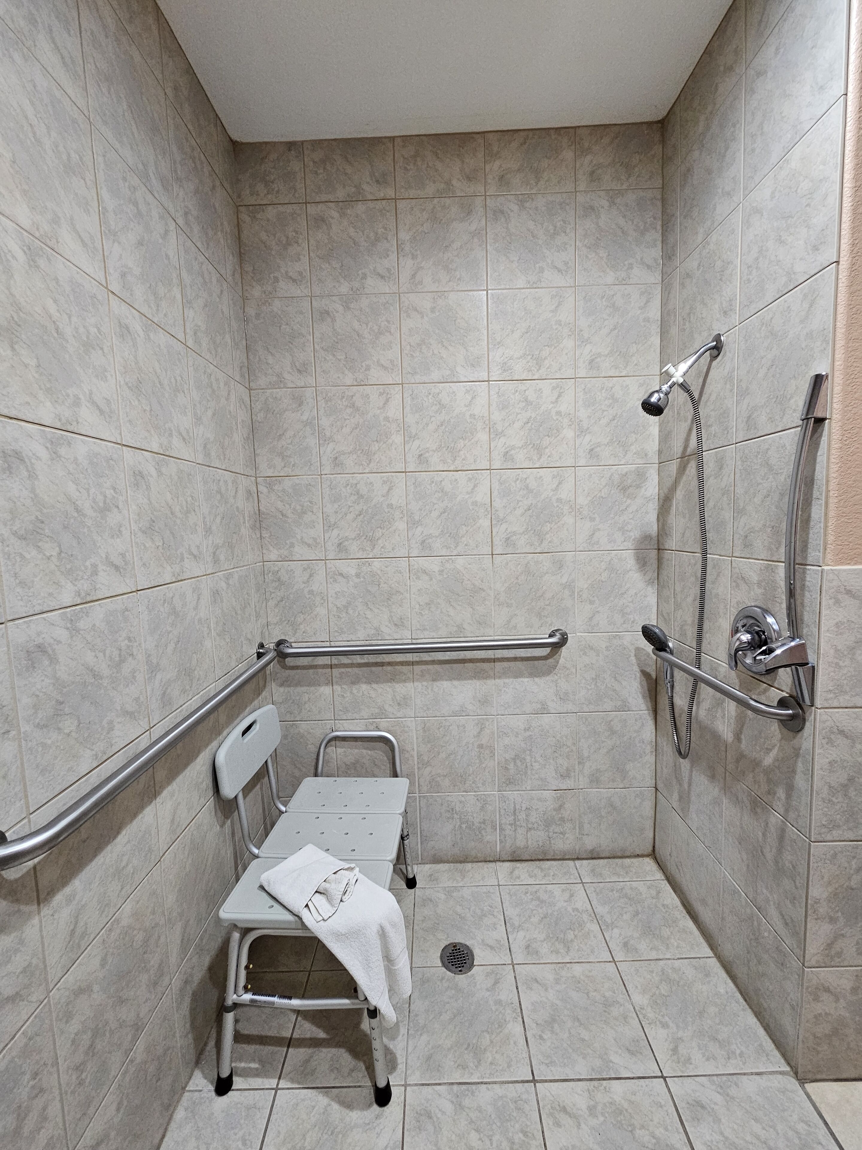 A small, tiled shower area with a grab bar, a wheelchair, and a handheld showerhead.