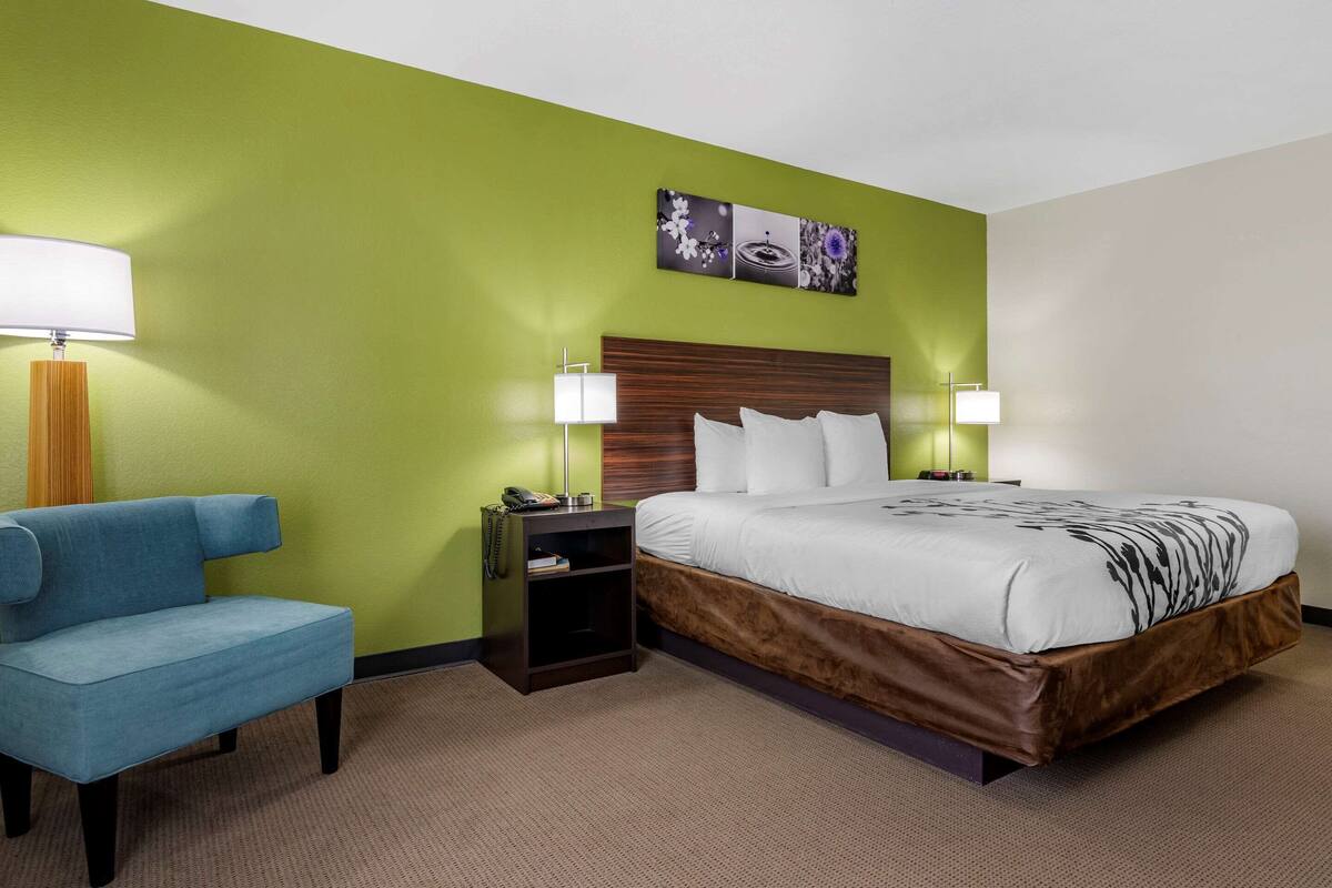 A hotel room with a bed, a blue chair, a nightstand with a lamp, and a wall with a floral artwork.