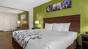 Quarto de hotel com uma cama grande, cabeceira de madeira, duas mesas de cabeceira com luminárias, uma cadeira e uma porta.