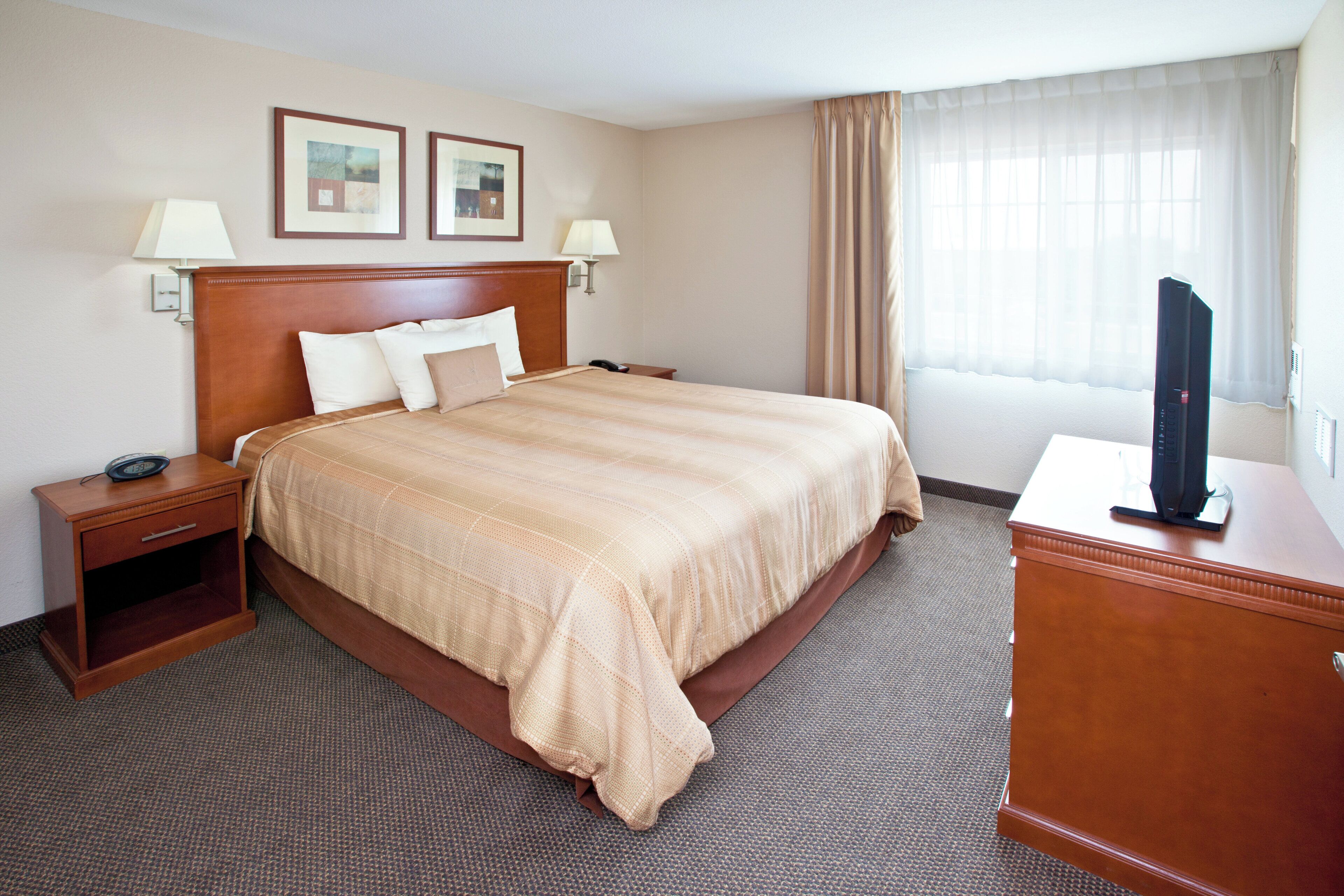 A hotel room with a large bed, two bedside tables, a TV on a stand, and a window with curtains.