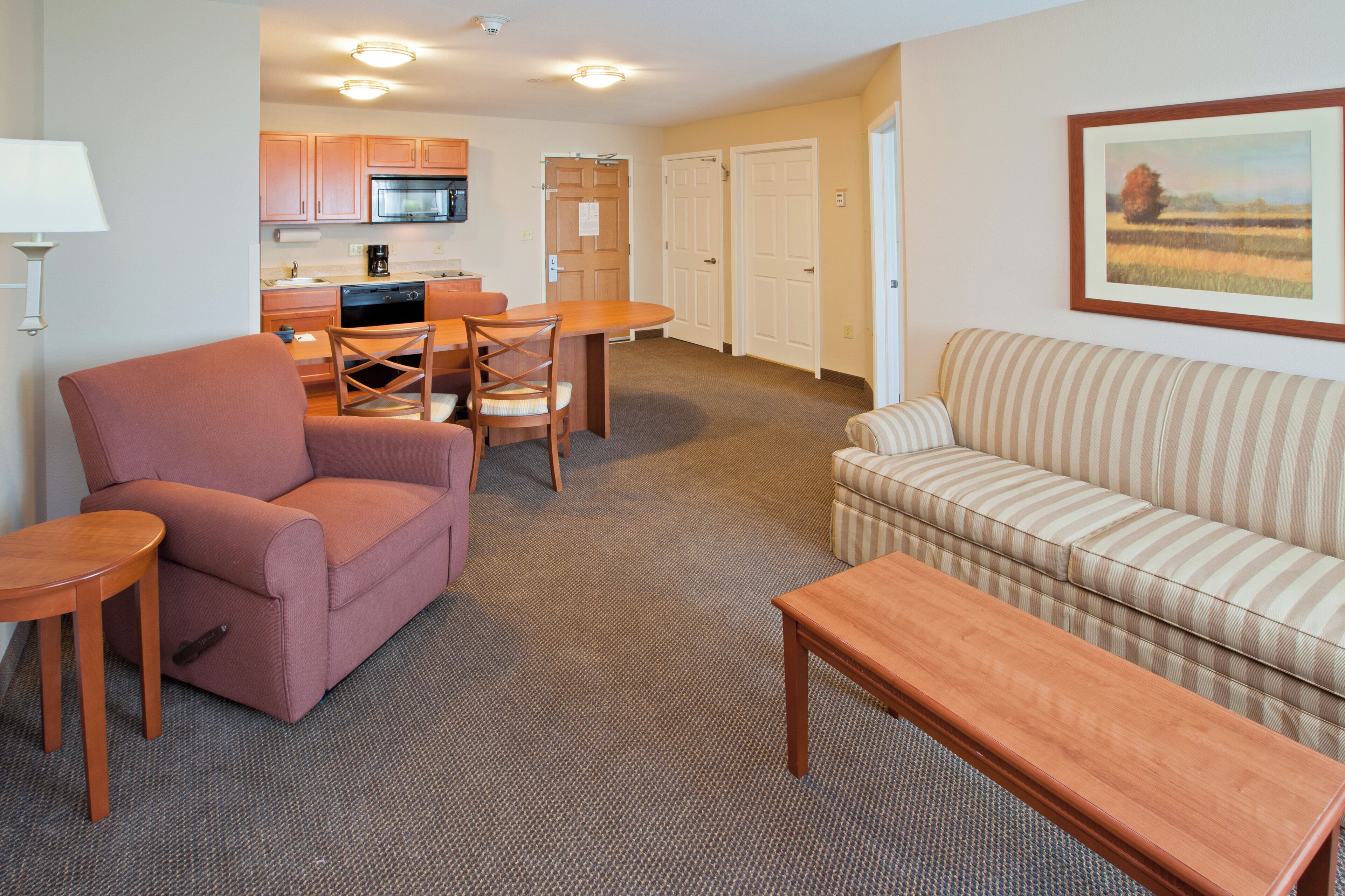 A living room with a sofa, armchair, and a wooden coffee table. There is a kitchen area with cabinets and a microwave.