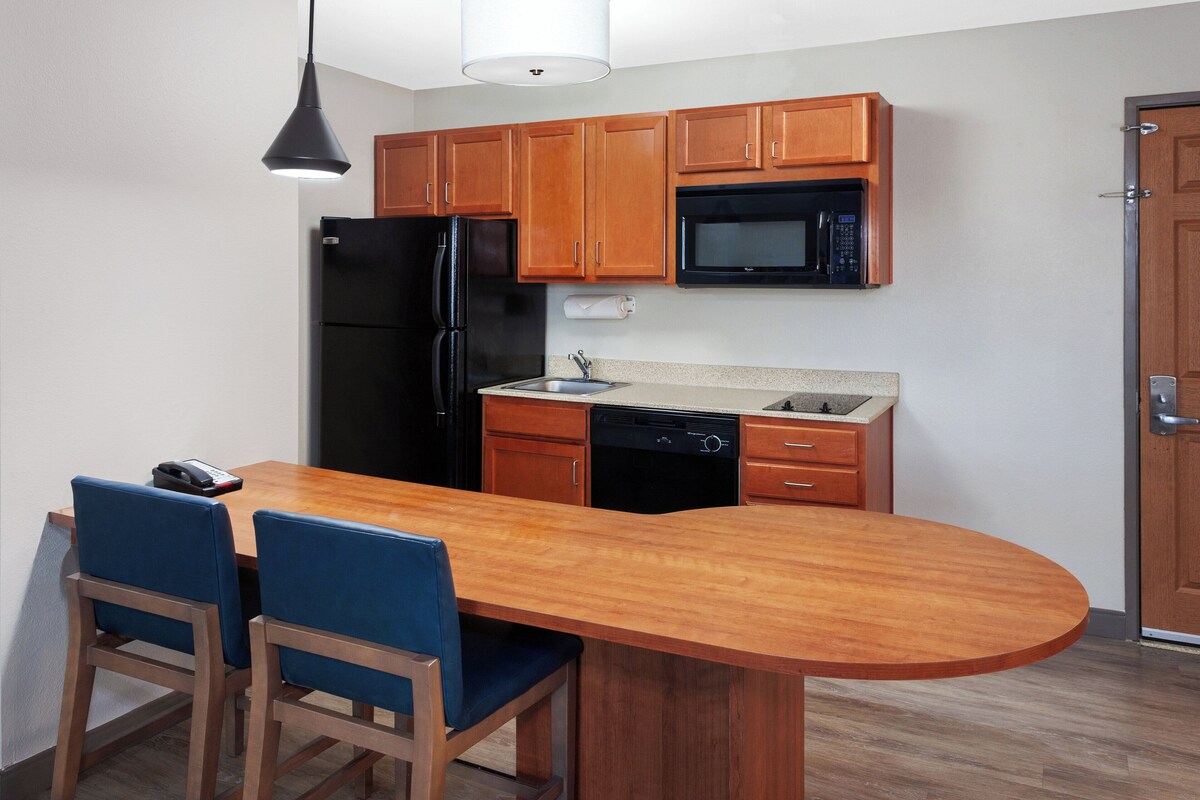 A compact kitchen with wooden cabinets, a black refrigerator, a microwave, and a dishwasher. There is a wooden island with two blue chairs.