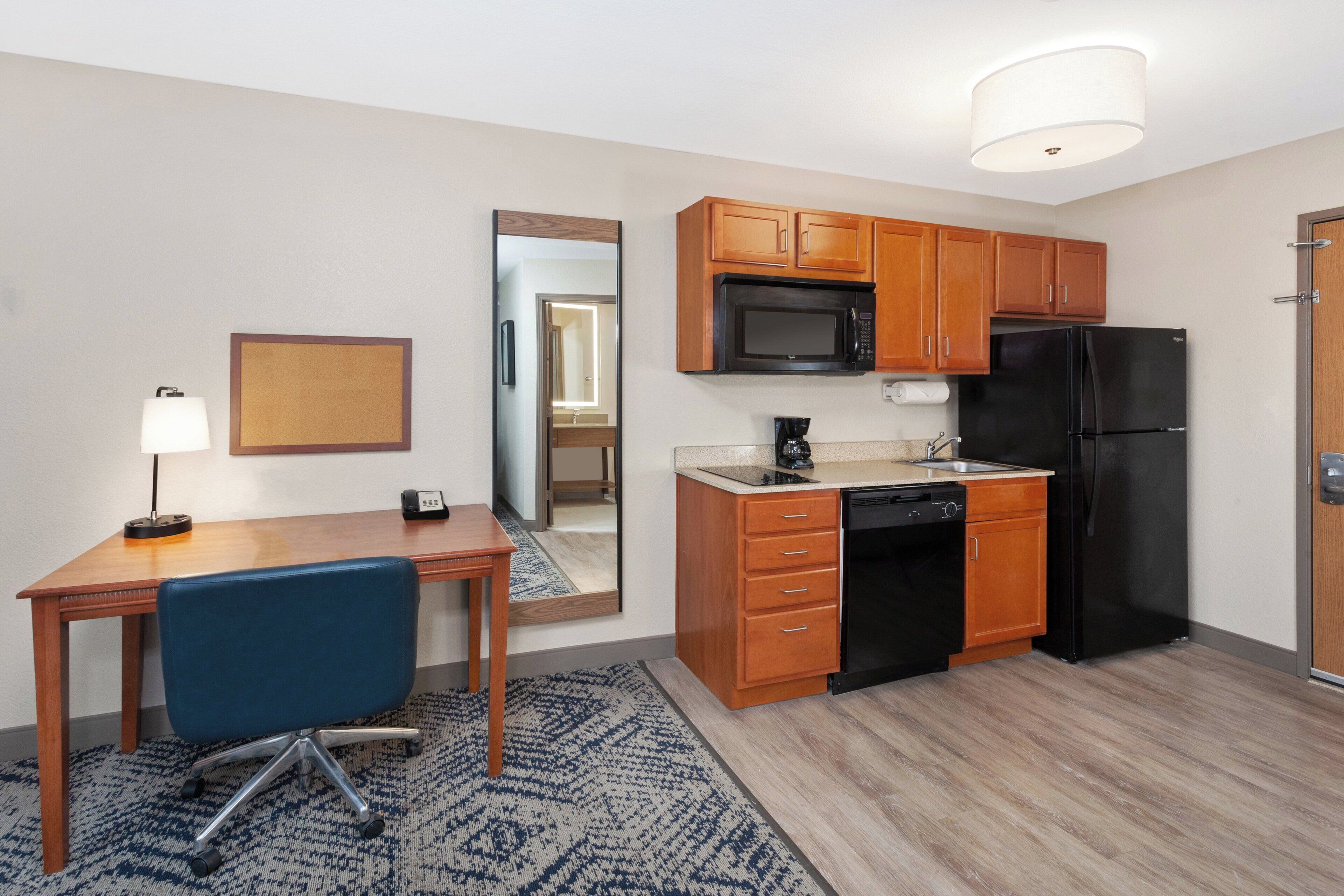 A compact hotel room with a kitchenette, a desk with a chair, a mirror, and a lamp.