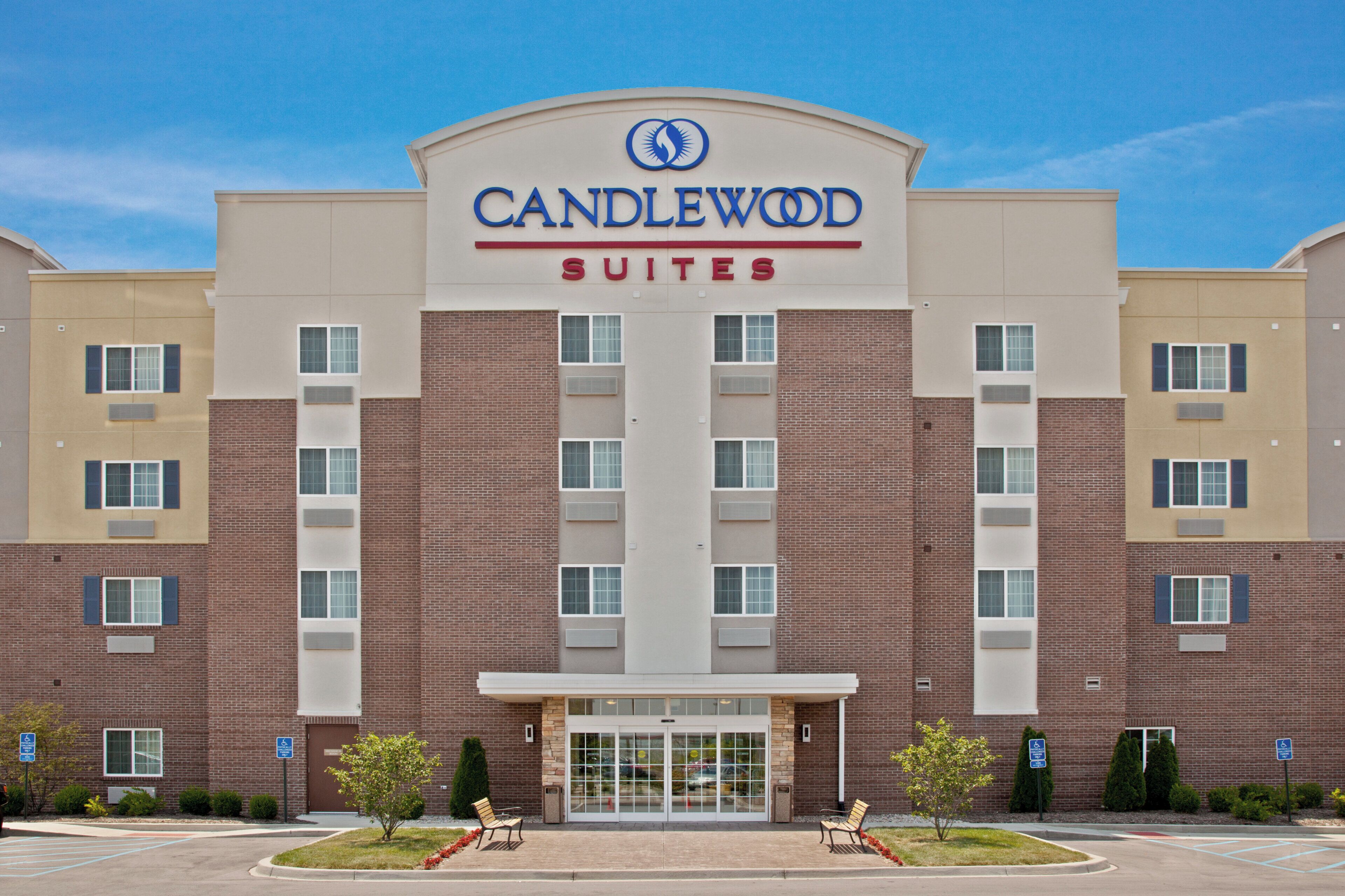 A hotel with a prominent 'CANDLEWOOD SUITES' sign, a brick facade, and a parking lot with a few trees and benches.