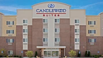 A hotel with a prominent 'CANDLEWOOD SUITES' sign, a brick facade, and a parking lot with a few trees and benches.