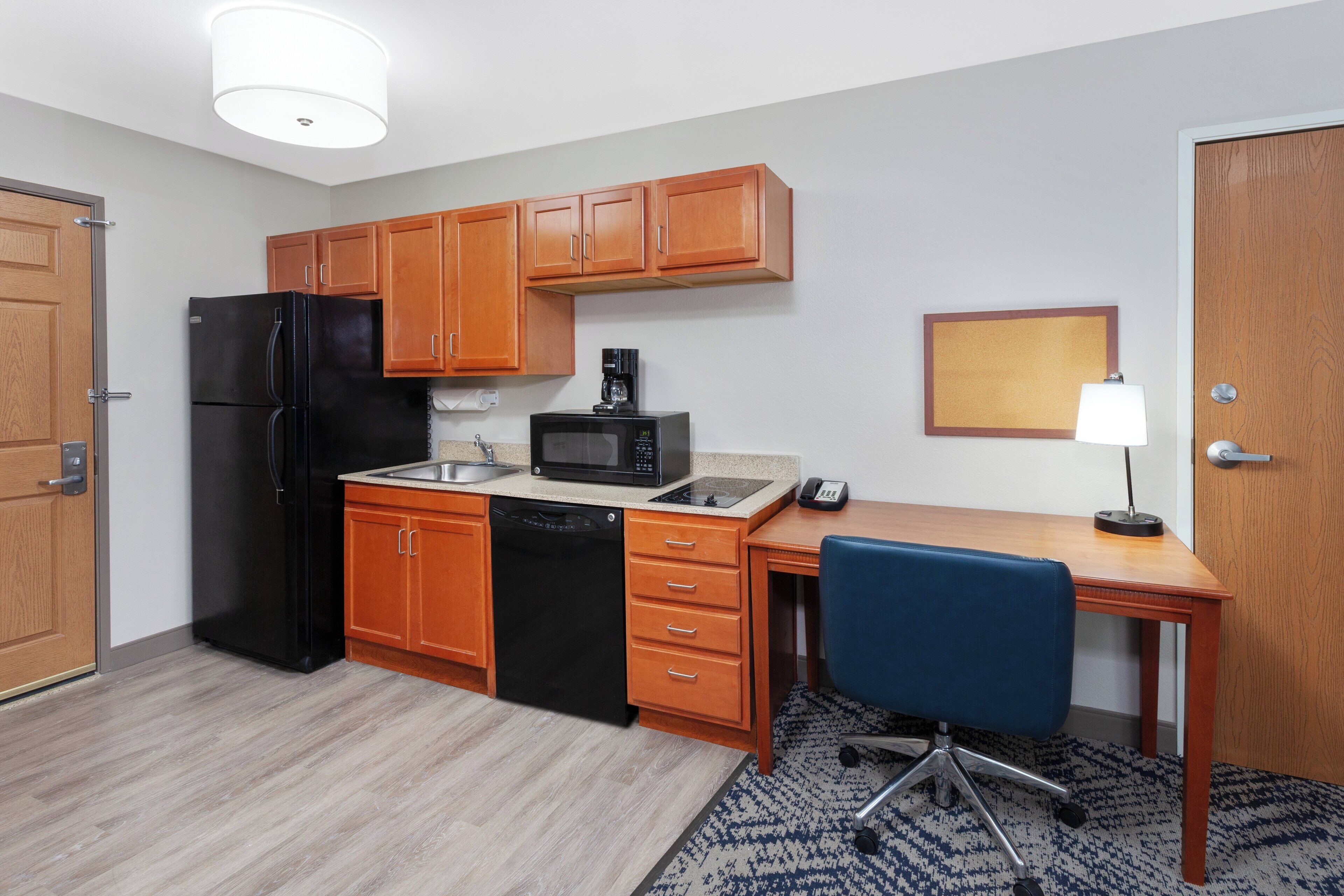 A compact kitchen with wooden cabinets, a black refrigerator, a microwave, and a small sink. There's a blue office chair and a desk with a lamp.