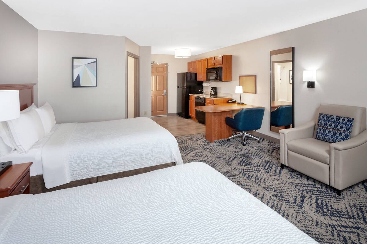 A hotel room with two beds, a desk, kitchenette, and a seating area.