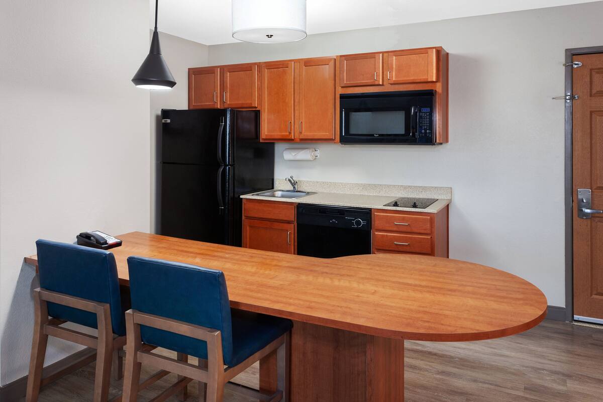 A compact kitchen with wooden cabinets, a black refrigerator, a microwave, and a dishwasher. There is a wooden island with two blue chairs.