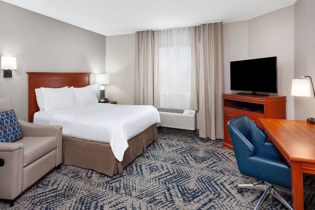 A hotel room with a large bed, a desk, a chair, a TV, and a window with curtains.