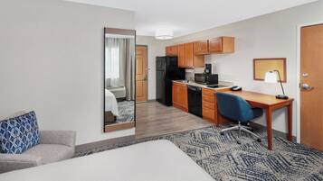 A hotel room with a bed, a desk, a chair, a kitchenette, and a mirror.