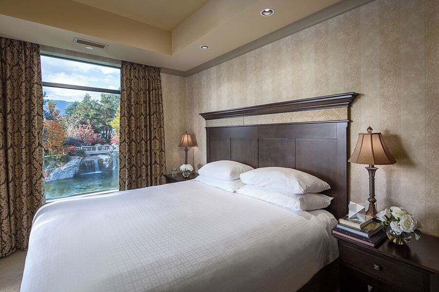 A hotel room with a large bed, two bedside lamps, a nightstand with books and flowers, and a view of a waterfall through a window.