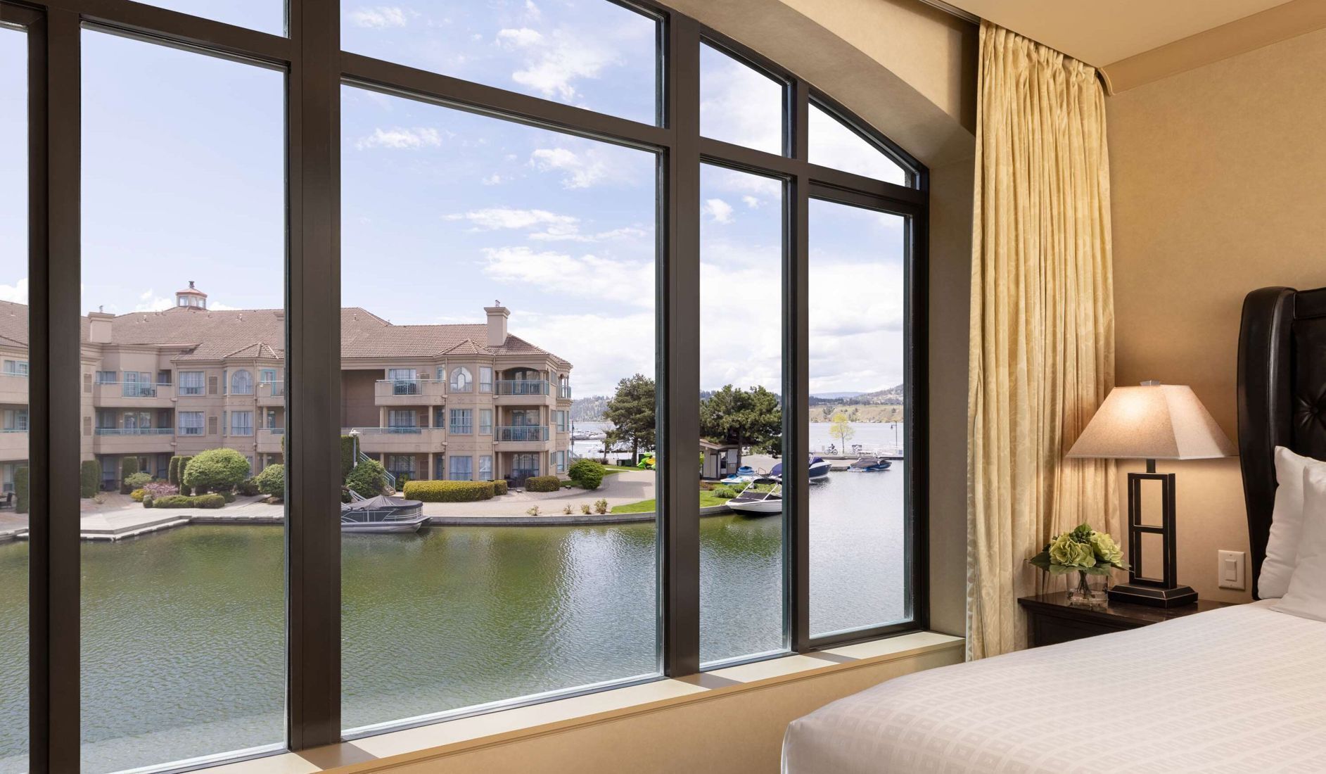 A hotel room with large windows, a bed, a nightstand, a lamp, and a view of a water body and buildings.