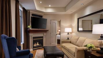 A hotel room with a fireplace, a television, a sofa, a chair, and a bedside table with a lamp.