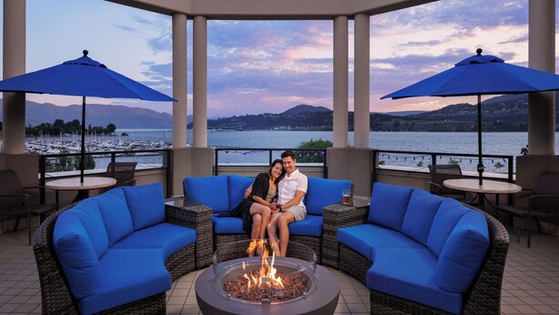 A couple seated on a blue sofa with a fire pit in the center, surrounded by outdoor tables and blue umbrellas, overlooking a lake.