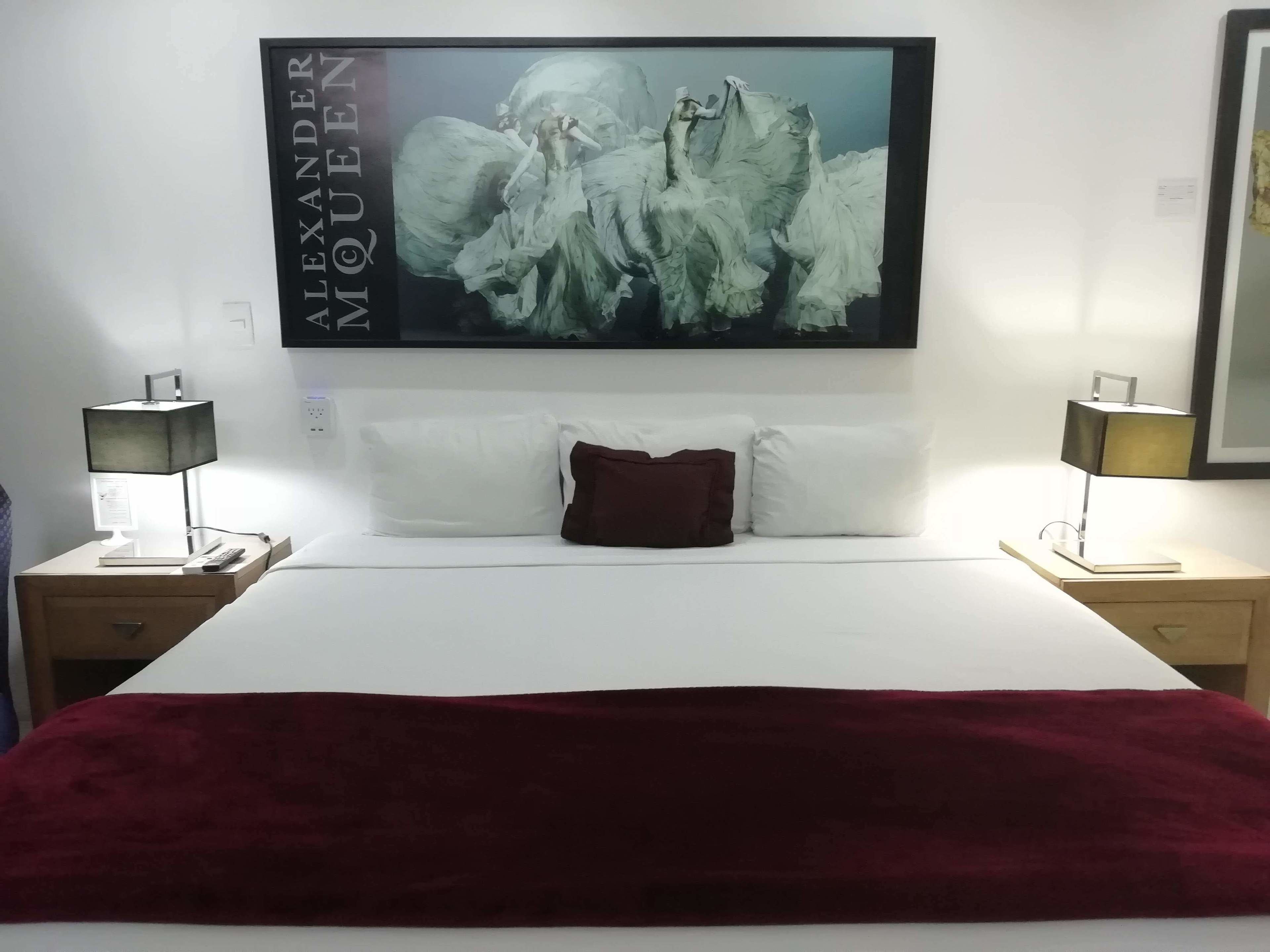 Una habitación de hotel con una cama grande, ropa de cama blanca, una manta de color rojo oscuro y una obra enmarcada sobre la cama. Hay dos mesitas de noche con lámparas y un buró con una lámpara y un libro.