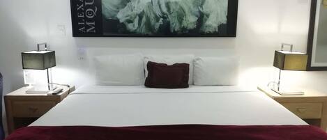 Una habitación de hotel con una cama grande, ropa de cama blanca, una manta de color rojo oscuro y una obra enmarcada sobre la cama. Hay dos mesitas de noche con lámparas y un buró con una lámpara y un libro.
