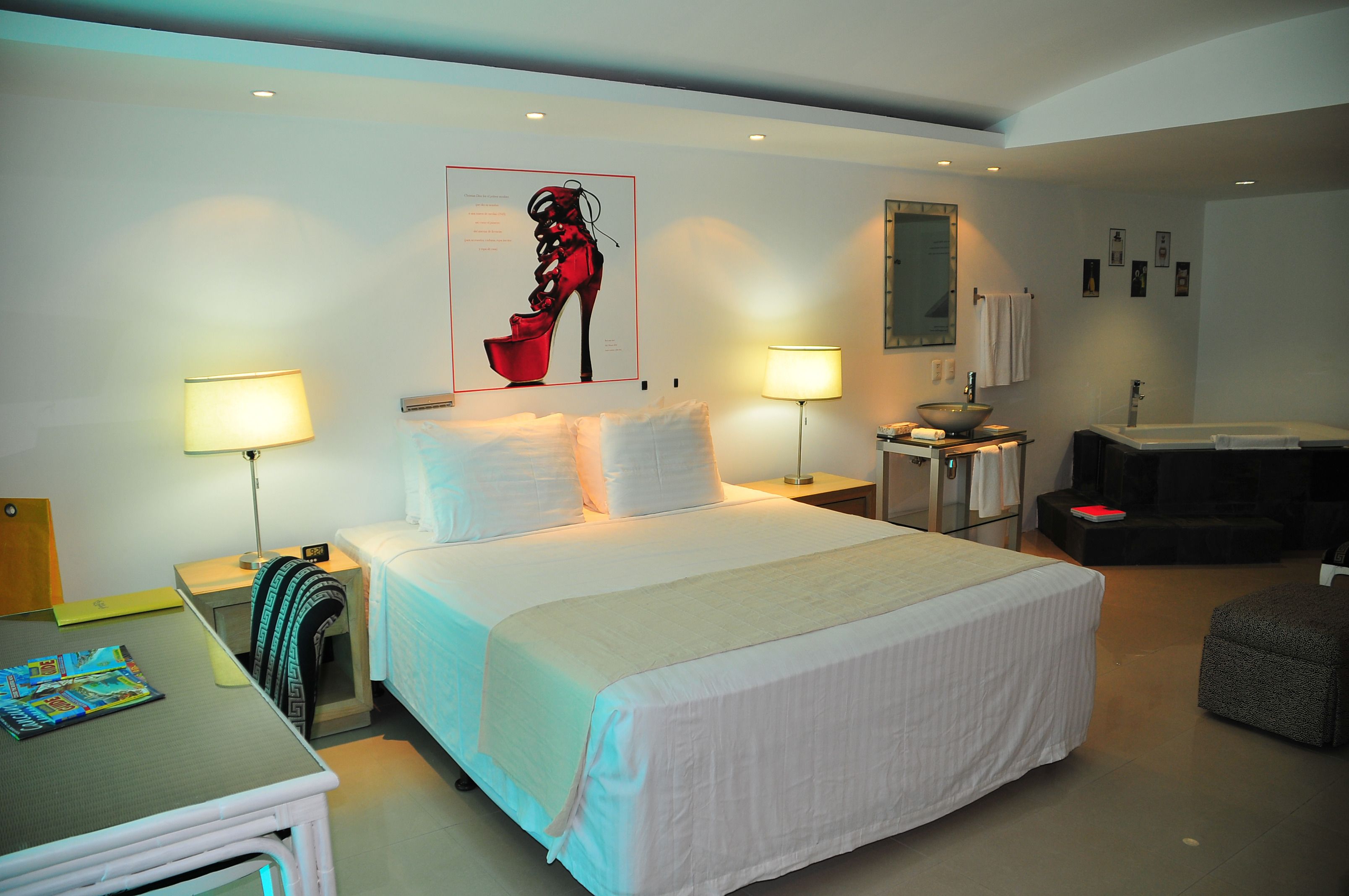 A hotel room with a large bed, two bedside tables with lamps, a desk with a chair, a small stool, and a bathroom with a bathtub and sink.