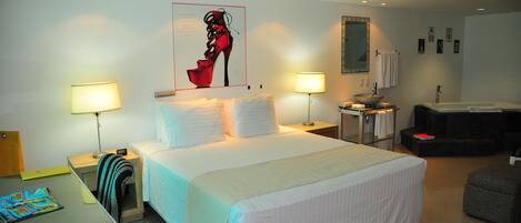 A hotel room with a large bed, two bedside tables with lamps, a desk with a chair, a small stool, and a bathroom with a bathtub and sink.