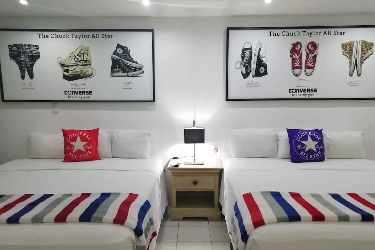 A hotel room with two beds, each with a pillow featuring the Converse All Star logo. There are framed pictures of Converse All Star sneakers on the wall above the beds.
