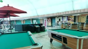 Un espace dédié aux tables de billard, avec deux tables de billard, un parasol rouge et un coin bar avec des tabourets et un comptoir.