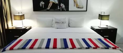 A hotel room with a large bed, two bedside tables with lamps, a framed picture of a model and a shoe, and pillows with the brand name.