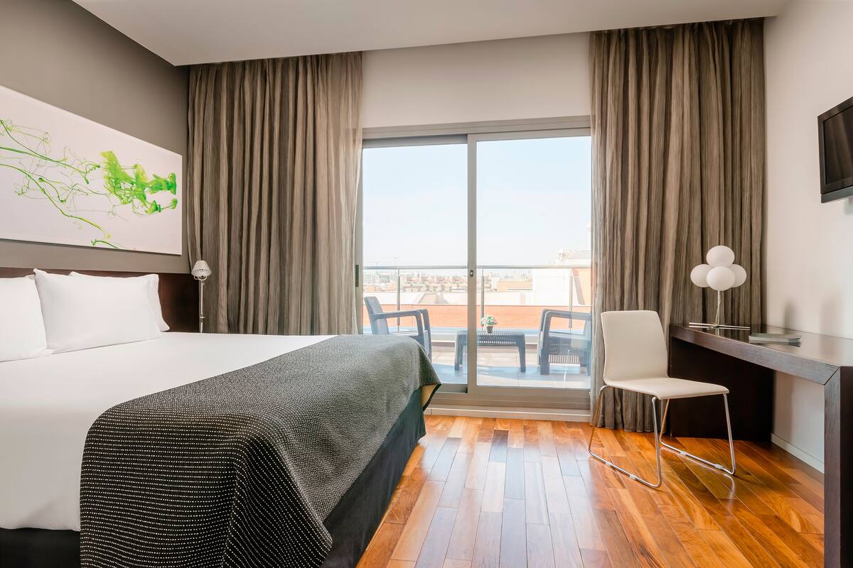 A hotel room with a large bed, a desk with a chair, a view through a sliding door, and a painting on the wall.