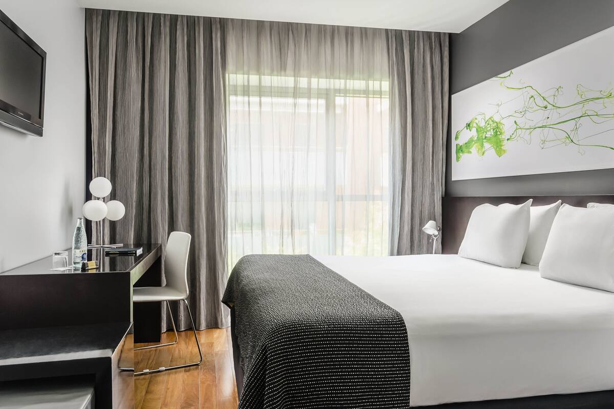 A modern hotel room with a large bed, a desk with a chair, a TV, and a large window with curtains.
