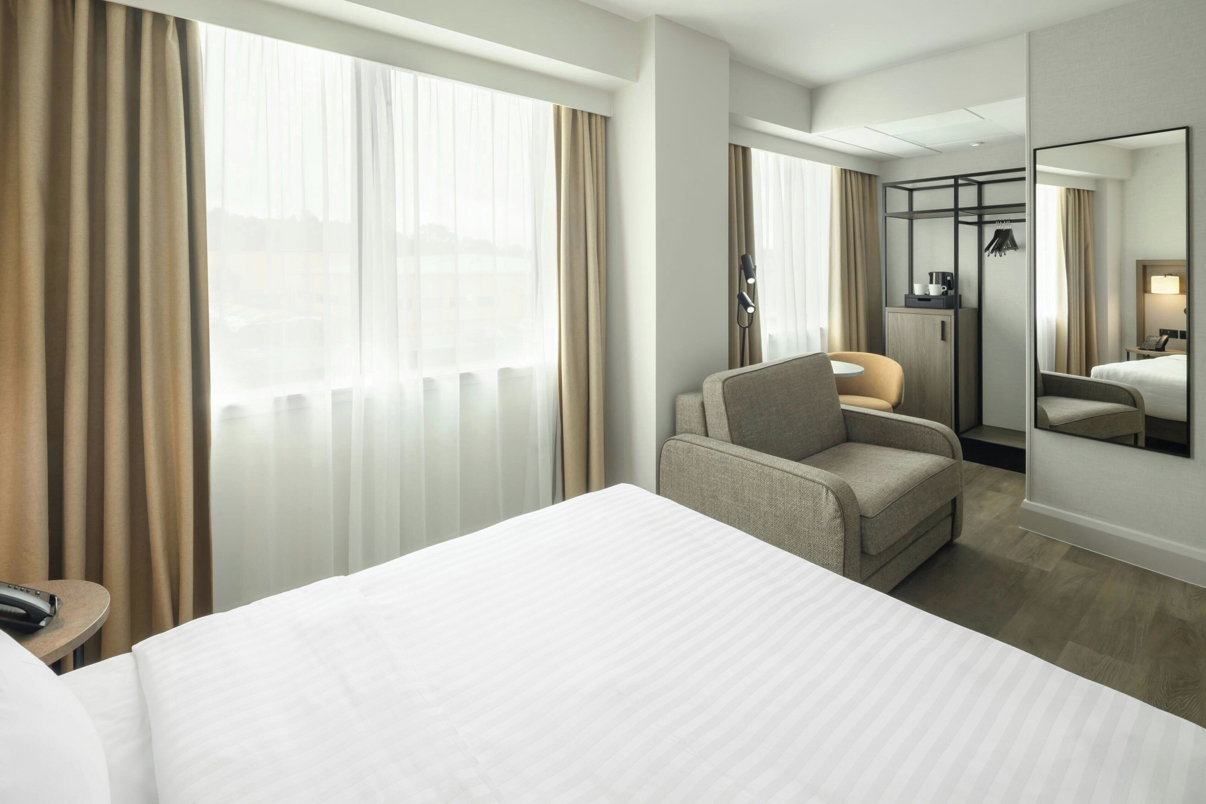 A hotel room with a bed, a chair, a desk, a mirror, and a window with curtains.