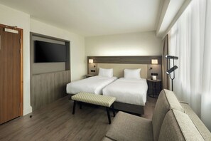 A hotel room with two beds, a TV, bedside lamps, a bench, and a sofa.